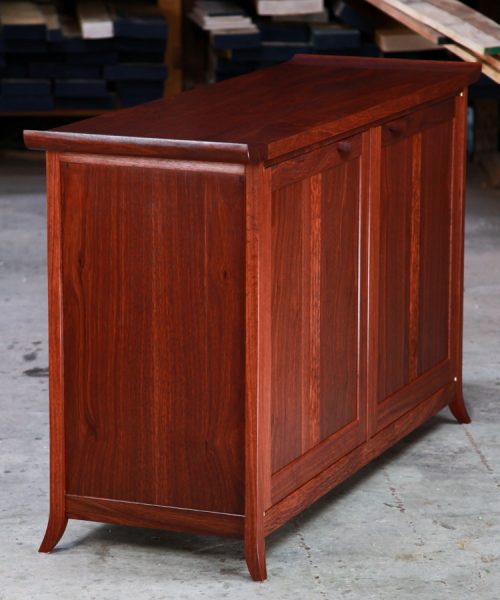 The side view of a beautiful jarrah cabinet sitting in a workshop in front of a timber rack.