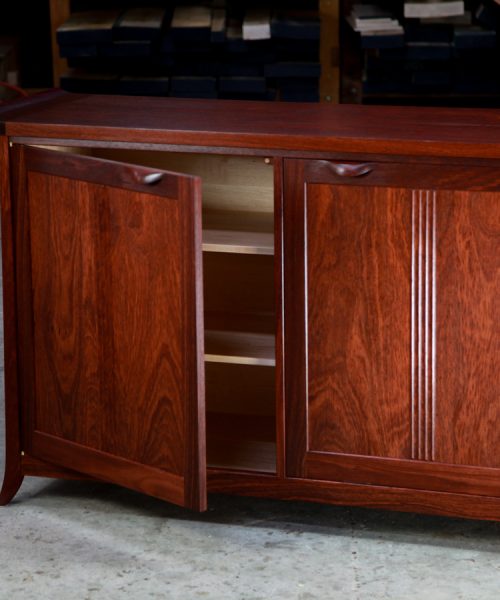 A single door is open of a beautiful jarrah cabinet sitting in a workshop in front of a timber rack.