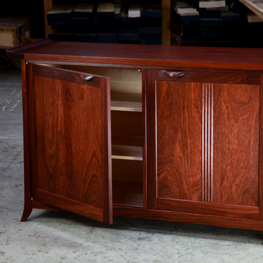 A single door is open of a beautiful jarrah cabinet sitting in a workshop in front of a timber rack.