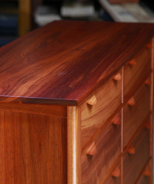 A beautiful jarrah cabinet with hand made drawer handles sits in a workshop in front of a timber rack.