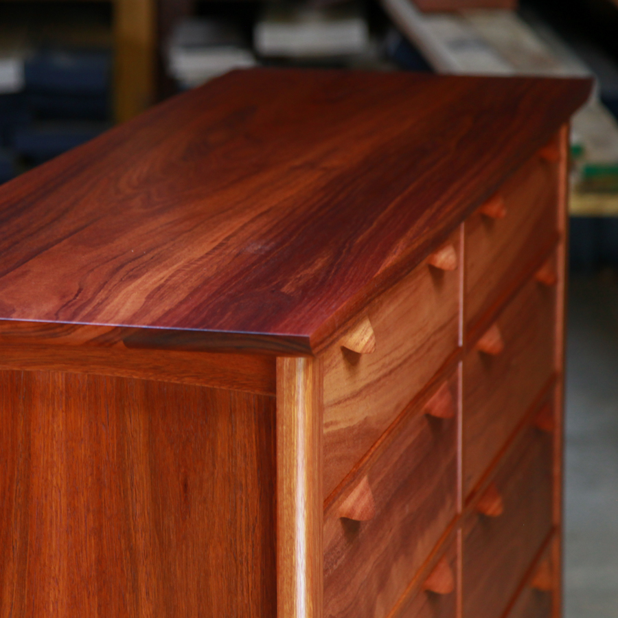 A beautiful jarrah cabinet with hand made drawer handles sits in a workshop in front of a timber rack.