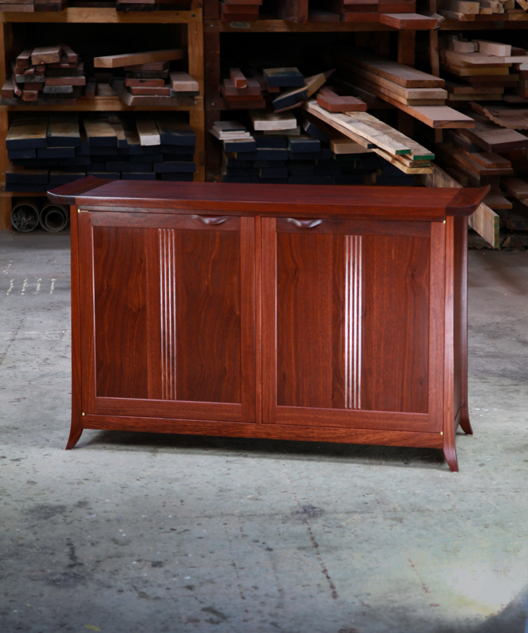 A beautiful jarrah cabinet sits in a workshop in front of a full timber rack.