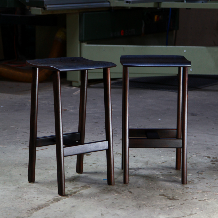 A moody photograph of two black timber stools in front of saw in a workshop.