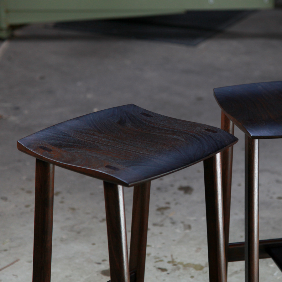 A moody photograph of two black timber stools in front of saw in a workshop.