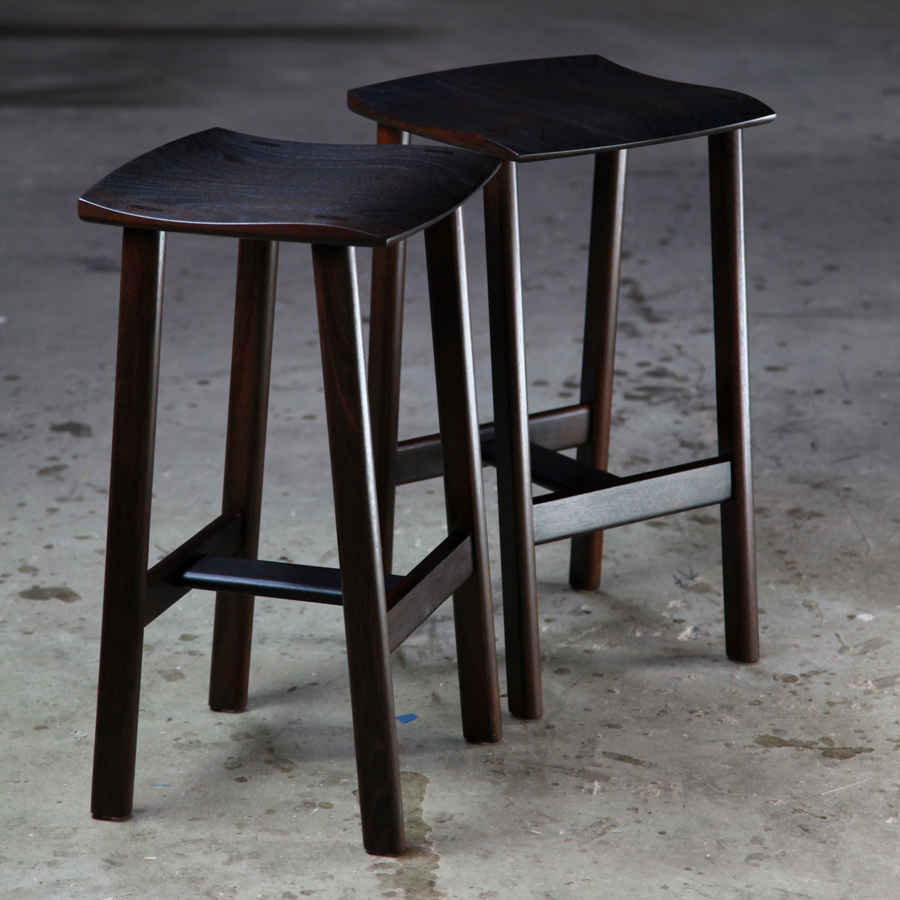 A moody photograph of two black timber stools in front of saw in a workshop.