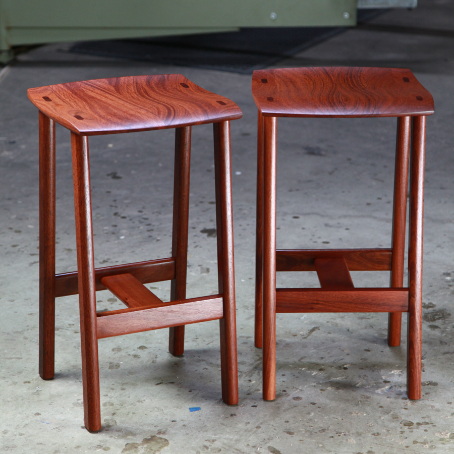 Two perfectly matched jarrah stools sit in a woodworking workshop.